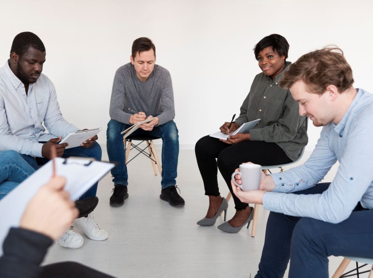 A diverse group of five adults sits in a circle, some holding notebooks or clipboards, and one person holding a mug, engaged in a thoughtful discussion in a bright, minimalistic room.