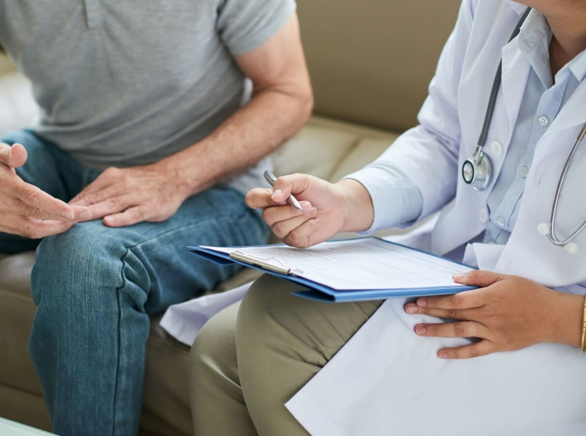 A doctor in a white coat takes notes on a clipboard while sitting next to a patient. The patient appears to be discussing something, with hands gesturing, while the doctor listens attentively.