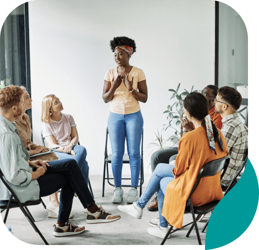 A woman stands and speaks to a diverse group of people seated in a circle, suggesting a group discussion or support session in a bright, modern room with plants in the background.