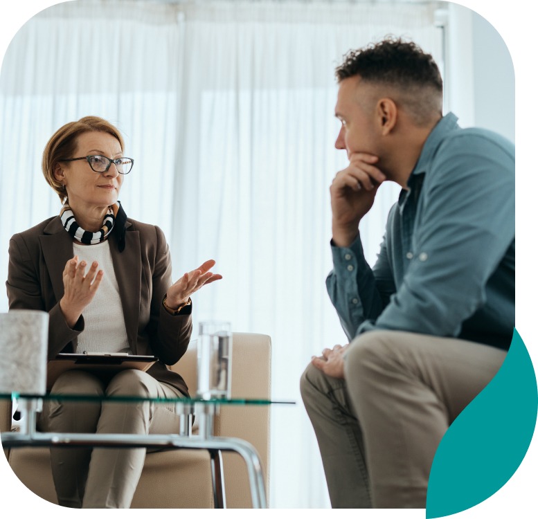 A woman and a man sit facing each other in a bright room. The woman gestures while holding a clipboard, and the man listens with his hand on his chin, suggesting a serious conversation or counseling session.