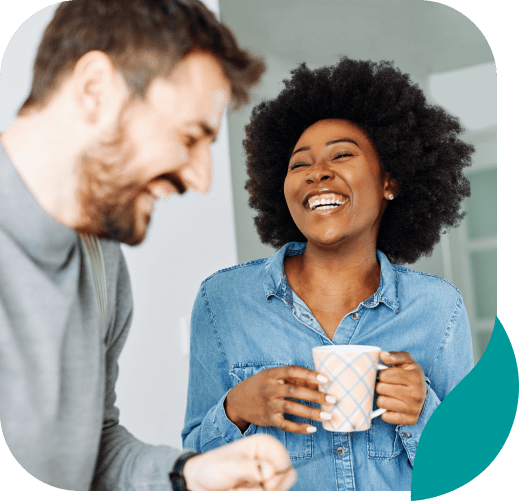 A woman in a denim shirt holding a mug smiles and laughs with a man who is also laughing, both appearing to enjoy a friendly conversation indoors.