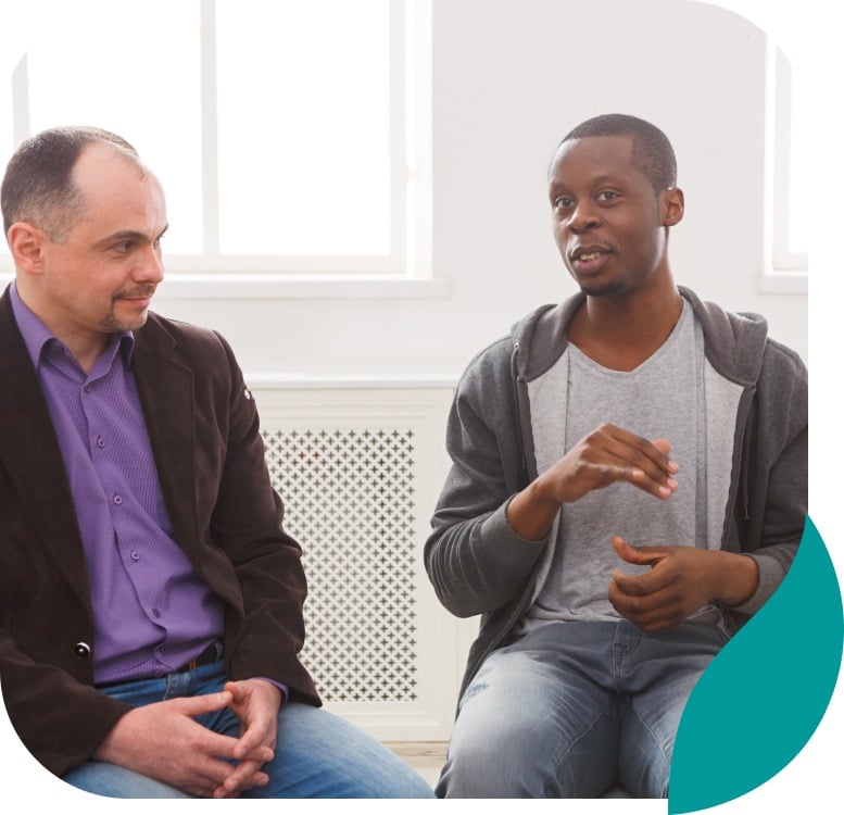 Two men sitting and talking in a brightly lit room; one man gestures with his hands while the other listens attentively. Both are casually dressed, and a large window is in the background.