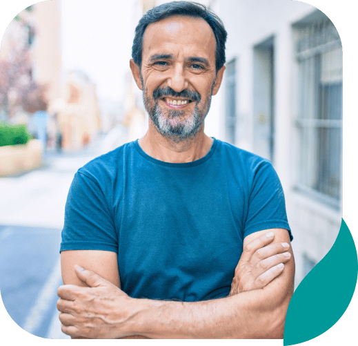 A middle-aged man with dark hair and a beard stands outside with his arms crossed, smiling confidently. He is wearing a blue t-shirt, and the background shows an urban street scene.