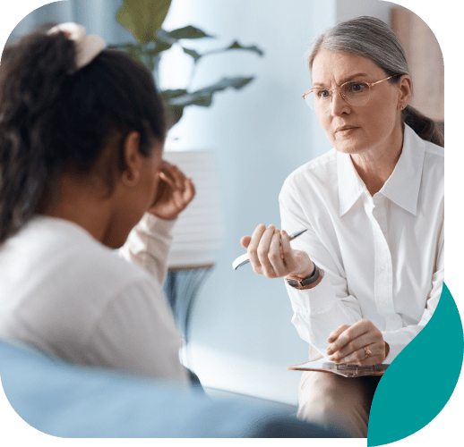 A middle-aged woman in glasses listens attentively to a younger woman who appears distressed, sitting with her head in her hand during a counseling or therapy session in a comfortable, softly lit room.