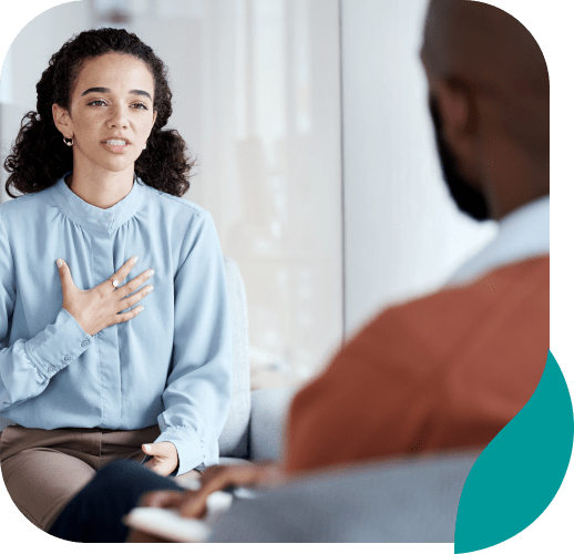 A woman sits across from a man, speaking with her hand on her chest in a thoughtful, expressive gesture during a conversation or counseling session.