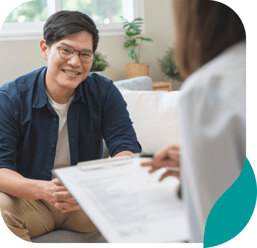 A man wearing glasses and a blue shirt sits on a couch, smiling and talking to another person who is holding a clipboard, suggesting a therapy or counseling session in a comfortable indoor setting.