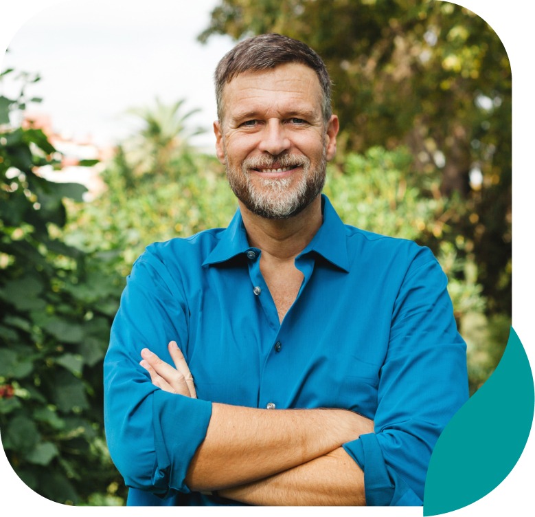 A middle-aged man with short hair and a beard, wearing a blue shirt, stands outdoors with arms crossed and smiles at the camera, surrounded by greenery.