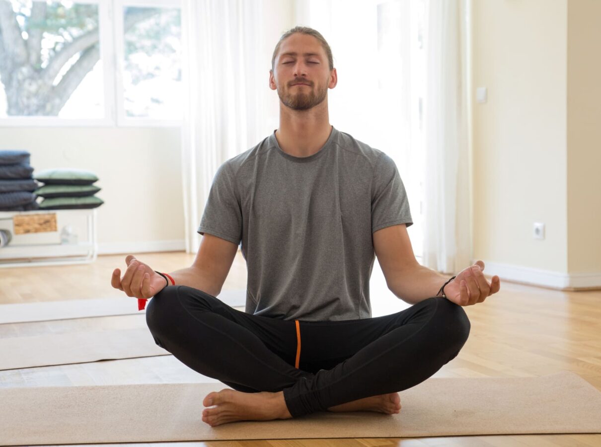 A man in a gray t-shirt and black pants sits cross-legged on a yoga mat indoors, eyes closed, meditating with hands resting on his knees in a bright room with wooden floors, highlighting how Mental Health Matters.