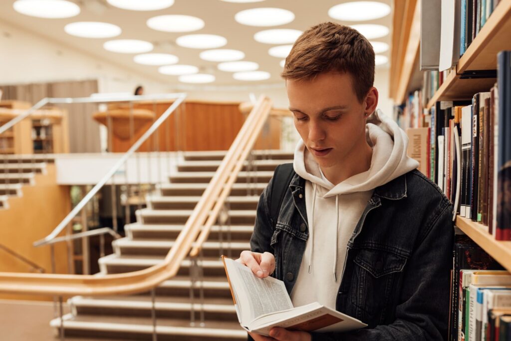 A young person in a denim jacket and hoodie reads a book next to bookshelves in a modern library, with wide stairs and circular ceiling lights in the background, embracing the idea that Mental Health Matters through the power of reading.