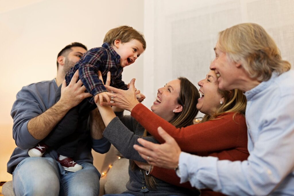 A joyful family plays together indoors, with an adult lifting a laughing young child as three other adults reach out, all smiling and enjoying the moment—showing how Mental Health Matters in creating happy memories together.