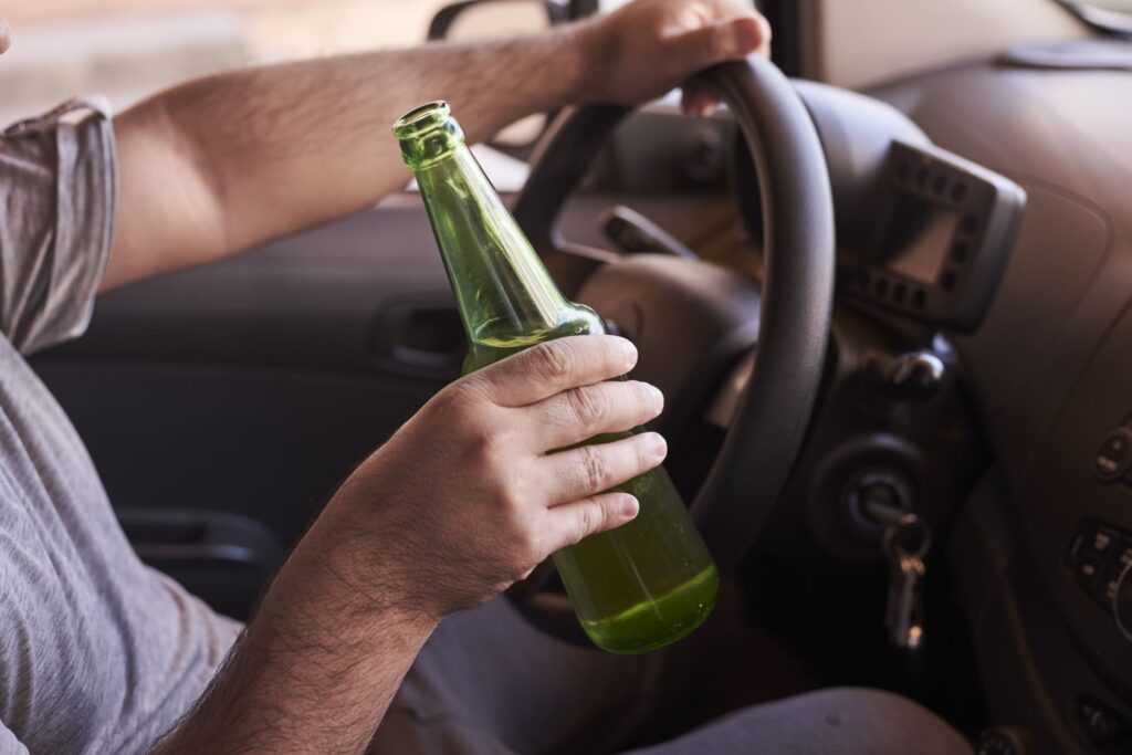 A person sits in the driver’s seat, gripping the steering wheel with one hand and holding a green glass bottle in the other. The car keys are in the ignition—a stark reminder that mental health matters behind the wheel.