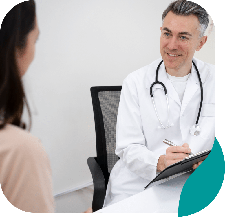 A doctor in a white coat with a stethoscope around his neck smiles while talking to a patient and taking notes on a clipboard in a medical office.