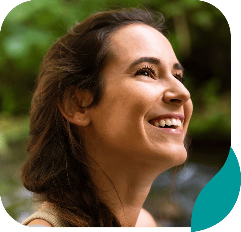 A young woman with long brown hair smiles and looks upward, with a blurred natural green background behind her.