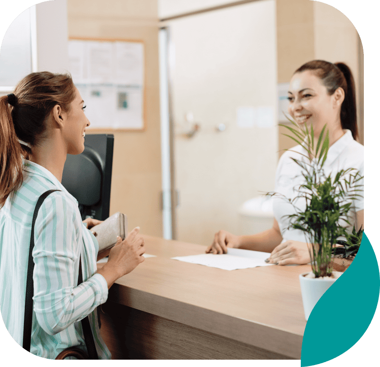 A woman stands at a reception desk, smiling and talking to a receptionist. The receptionist is seated behind the desk, which has a potted plant on it. Both women appear friendly and engaged in conversation.