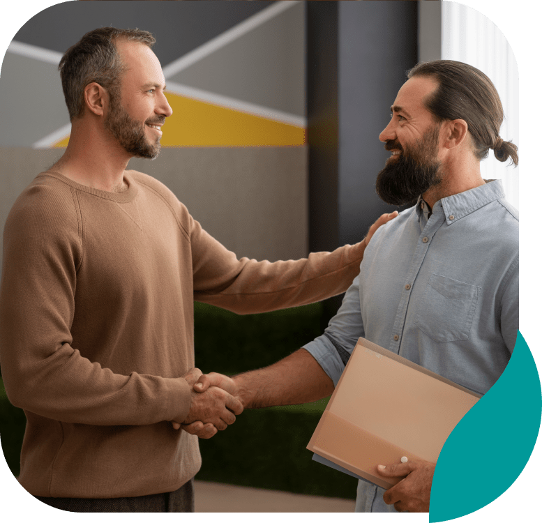 Two men smiling and shaking hands indoors; one man holds a folder under his arm while the other has a hand on his shoulder. They appear to be congratulating or greeting each other.