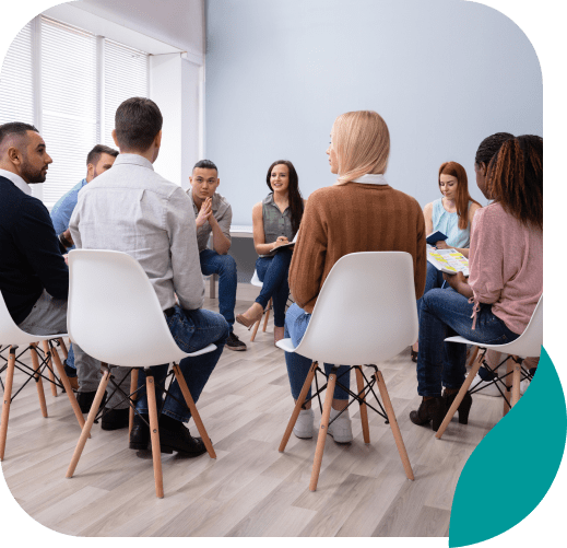 A group of people sits in a circle on white chairs in a bright room, engaged in a group discussion or support meeting.