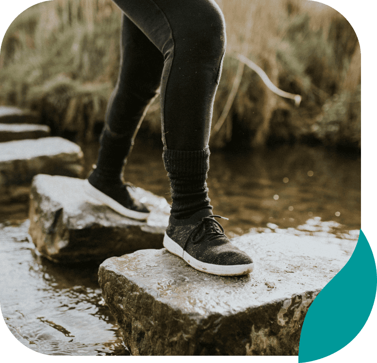 A person wearing black leggings and sneakers steps carefully across large stones in a shallow stream, surrounded by tall grass.