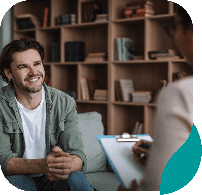 A man sits on a couch smiling and talking to a person holding a clipboard, suggesting a therapy or counseling session in a cozy room with bookshelves in the background.