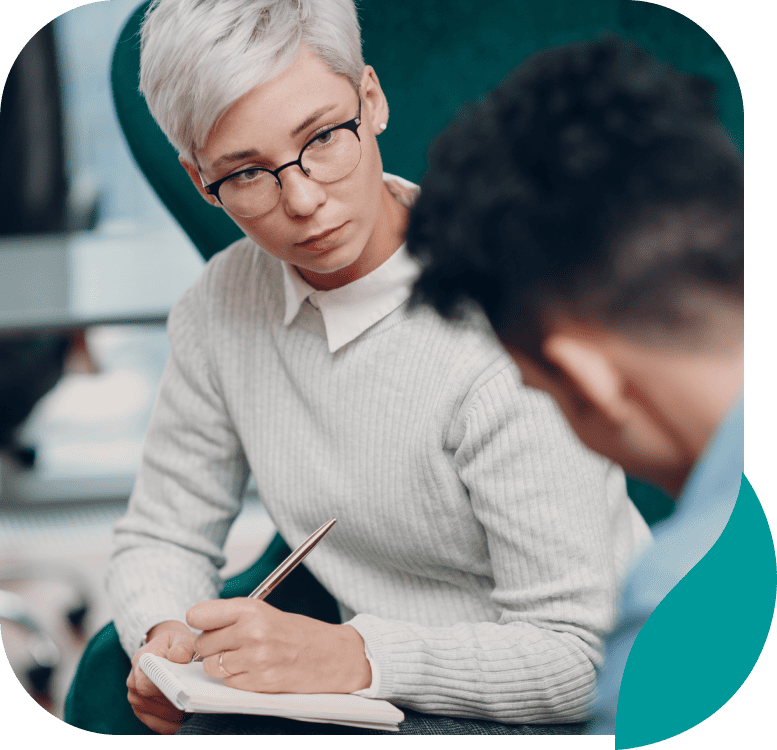 A person with short, light hair and glasses is sitting and taking notes on a pad while attentively listening to another person, suggesting a counseling or interview session.