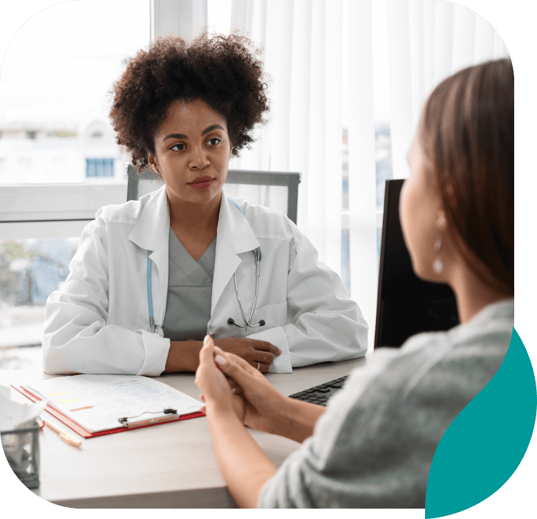 A female doctor with curly hair listens attentively to a patient across a desk in a bright medical office. The doctor has a stethoscope and notes in front of her, while the patient sits with hands clasped.