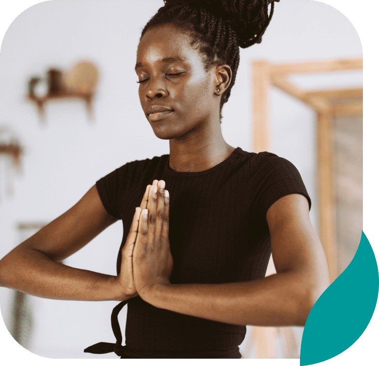 A woman with braided hair closed her eyes and pressed her palms together in a prayer position, appearing calm and focused while practicing yoga or meditation indoors.