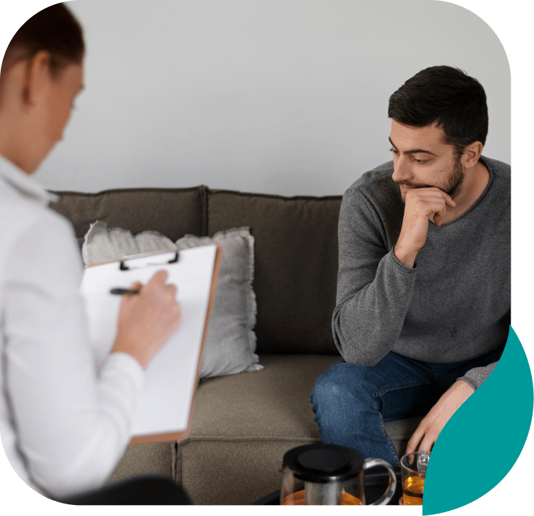 A man sits on a couch looking thoughtful while a person with a clipboard, possibly a therapist, takes notes during a session. A glass teapot and cup are on the table in front of them.