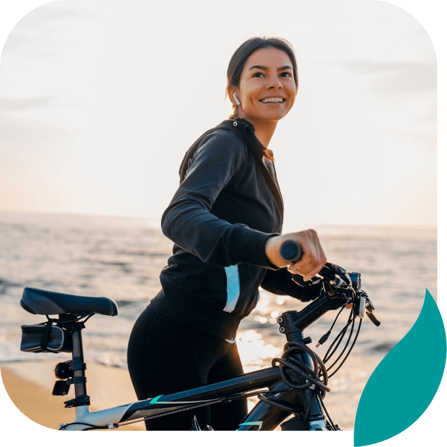 A woman in athletic wear smiles while standing beside her bicycle on a beach at sunset, with the ocean in the background.