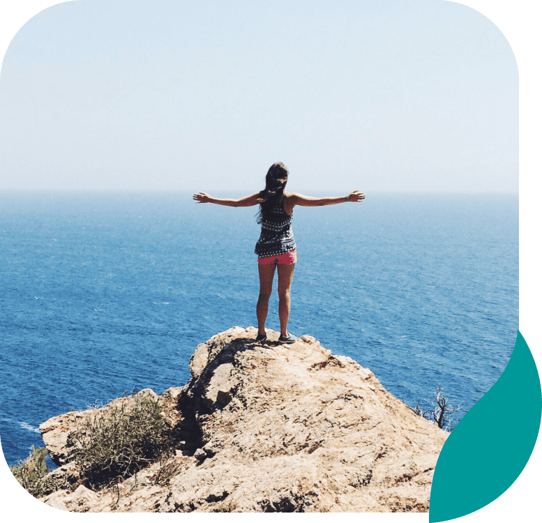 A person stands on a rocky cliff with arms outstretched, facing a vast blue ocean under a clear sky. Some shrubs are visible in the foreground. The image has rounded corners and a teal graphic in one corner.