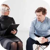 A woman with short blond hair and glasses holds a clipboard and talks to a man sitting beside her. The man, wearing a light blue shirt, looks down with a serious expression, hands clasped.