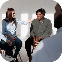 Four people are sitting in a circle having a discussion. One woman comforts another by placing a hand on her shoulder. The environment looks supportive and relaxed, with soft natural light.
