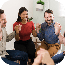 A group of people sitting in a circle indoors, holding hands and smiling, appearing to participate in a group support or therapy session.