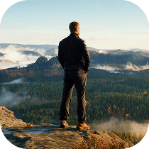 A man stands on a rocky cliff edge, looking out over a vast forest landscape with hills and scattered clouds or mist in the distance under a clear sky.