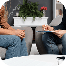 Two people sit across from each other in chairs; one holds a clipboard and pen, taking notes, while the other has hands clasped. A table and plants are in the background, suggesting a counseling or therapy session.