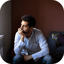 A man with dark curly hair and a beard sits indoors, resting his chin on his hand and looking thoughtfully out of a window. He wears a light blue shirt and appears deep in thought.