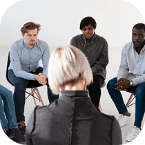 A group of four people sits in a circle listening to a person with short blonde hair, who is facing away from the camera, suggesting a group discussion or support meeting.
