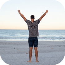 A person stands barefoot on a sandy beach facing the ocean, arms raised in celebration or joy, under a clear sky.