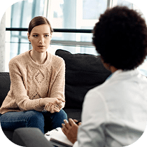 A woman in a light sweater sits on a couch with her arms crossed, listening attentively to another person holding a clipboard, suggesting a counseling or therapy session in a bright, modern office.