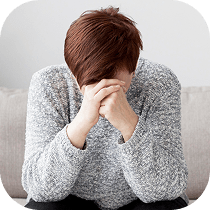 A person with brown hair sits on a couch, leaning forward with their elbows on their knees and their hands covering their face, appearing stressed or upset.