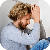 A man with curly blond hair and a beard sits against a wall, eyes closed, resting his forehead on his clasped hands, appearing stressed or deep in thought. He wears a gray sweatshirt and blue jeans.