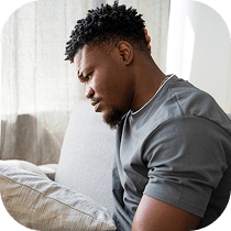 A man sits on a couch, leaning forward and holding his stomach with a pained expression, suggesting he may be experiencing discomfort or pain.