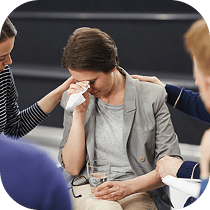 A woman sits holding a tissue and a glass of water, appearing upset and wiping her eyes, while two people offer comfort by touching her shoulder and arm.