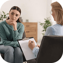 Two women sit facing each other in an office; one looks thoughtful with her hand on her chin, while the other, holding a clipboard and pen, appears to be taking notes during a conversation or counseling session.