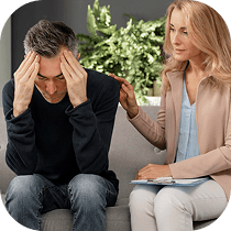A concerned woman sits beside a distressed man holding his head in his hands, gently comforting him with a hand on his shoulder. They are seated on a couch with plants in the background.
