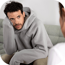 A man in a gray hoodie sits on a couch, leaning his head on his hand and looking thoughtfully at another person, whose back is visible in the foreground.