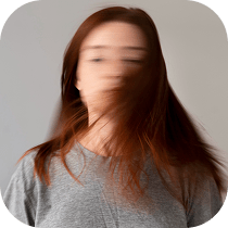 A person with long brown hair wearing a gray shirt, head blurred in motion, standing against a plain light background.