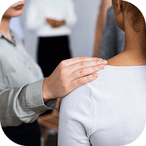 A person gently places their hand on another person's shoulder, offering comfort or support. Other people stand blurred in the background.