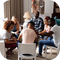 A diverse group of six people sit in a circle on chairs in a cozy living room, engaging in a supportive and empathetic group discussion. One person comforts another by touching their shoulder.