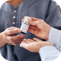 Close-up of one person handing a small white medicine bottle to another person, both wearing long-sleeved shirts. The background is softly blurred.