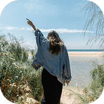 A person with long hair, wearing a denim jacket and black pants, walks toward a beach, surrounded by greenery, with one arm raised and pointing towards the blue sky and ocean.