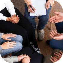 A group of people sitting in a circle with hands visible, some holding hands, and one person holding a clipboard and pen, suggesting a support group or discussion meeting.